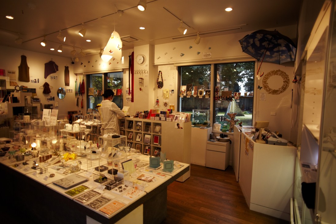 Interior of a museum shop with displayed merchandise, shelving, and large windows overlooking a garden.