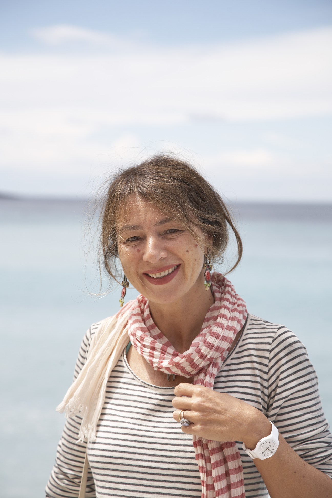 Rosanna Massarenti, a smiling woman with brown hair wearing a striped shirt and colorful scarf, standing against a blurred seascape background.