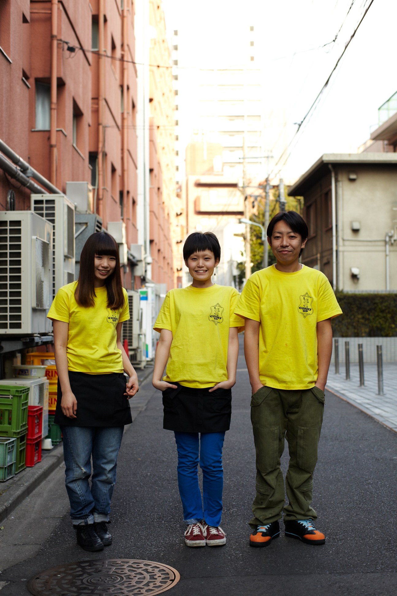 Three people in bright yellow Curry Up branded t-shirts stand in a narrow Tokyo alley lined with residential buildings.