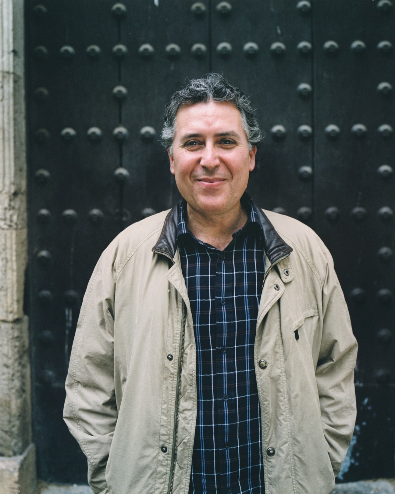 Man in beige jacket and plaid shirt standing against dark studded door.