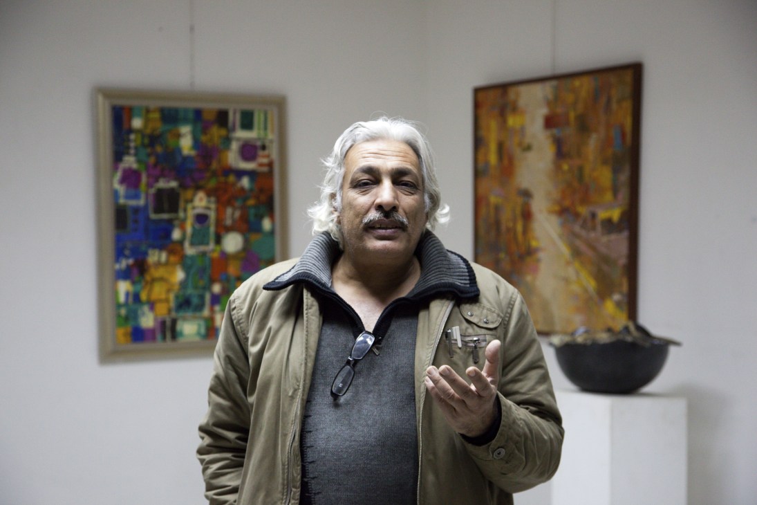 Iraqi artist Qasim Sabti with silver hair gestures while speaking in an art gallery.