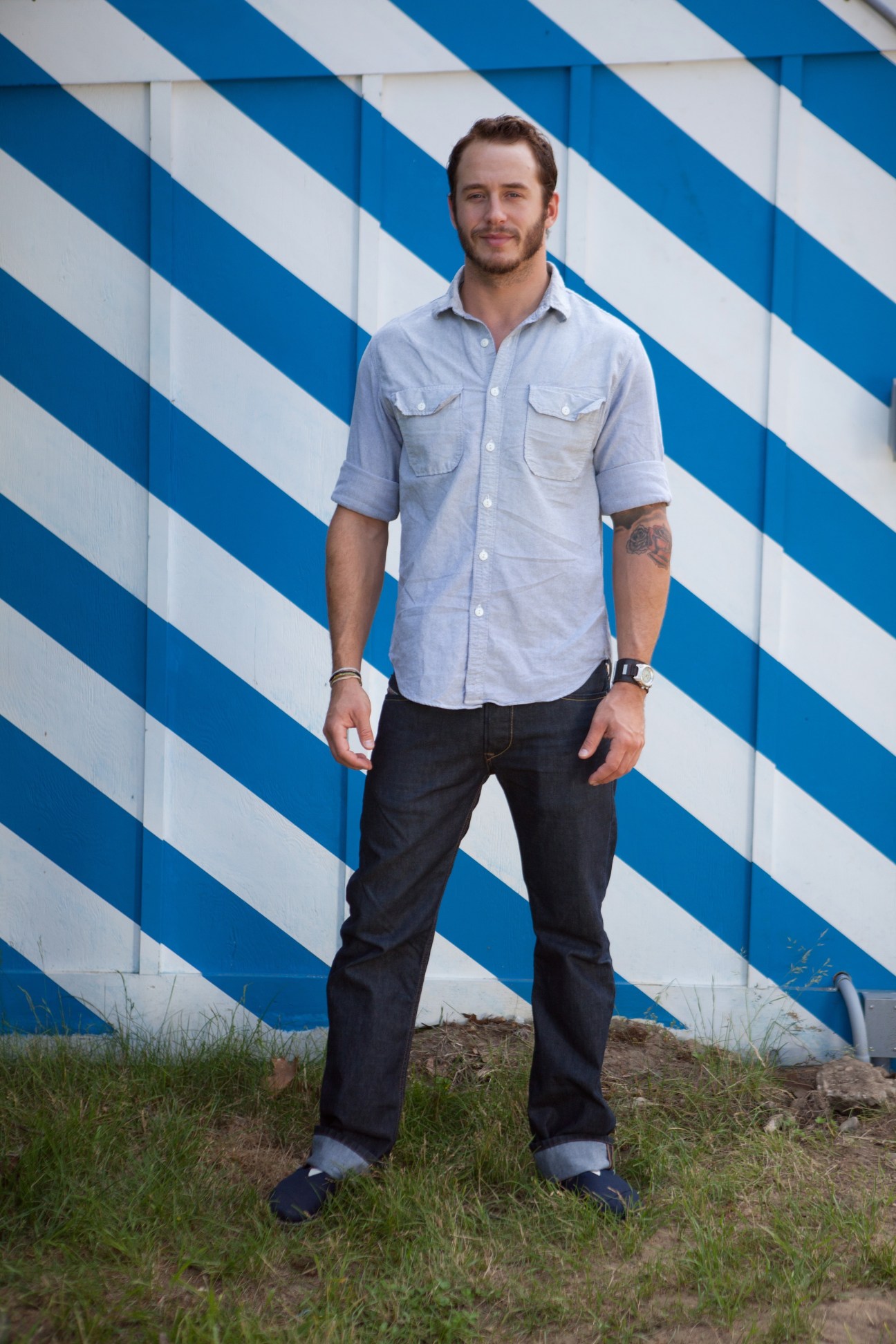 Man in light blue shirt and dark jeans standing in front of blue and white striped wall.
