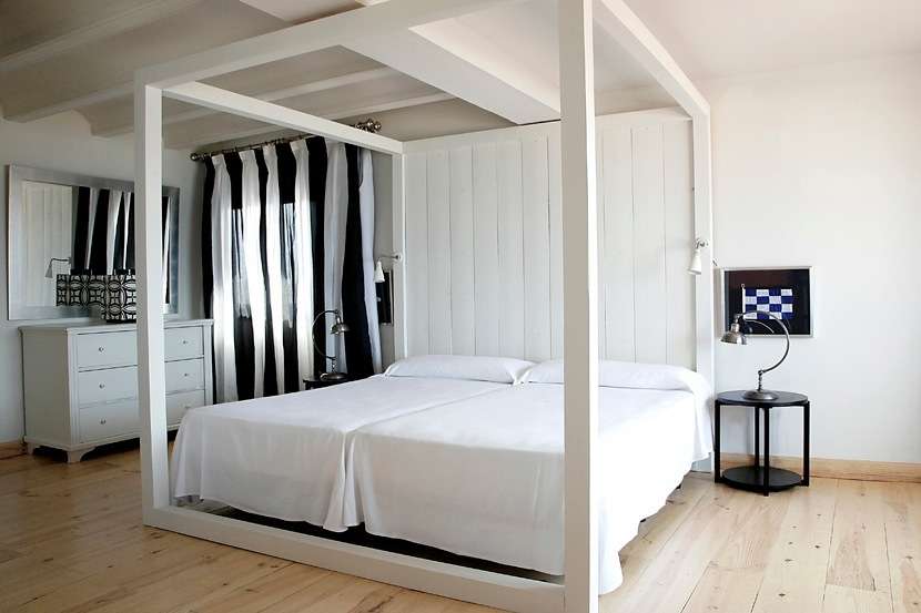 Modern bedroom with white four-poster bed, black and white striped curtains, wooden floor and minimalist furnishings.