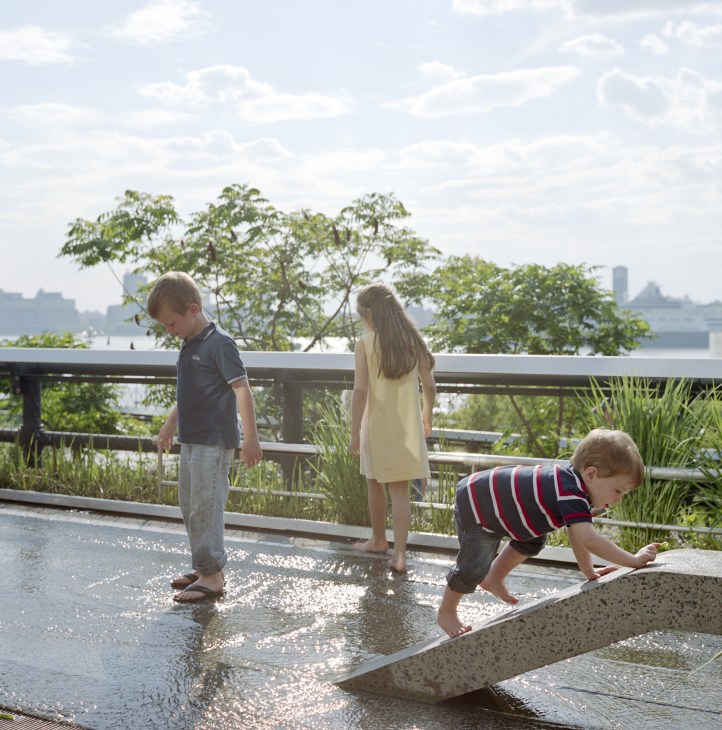 Three children play in shallow water on the High Line park with Manhattan skyline visible in the background.