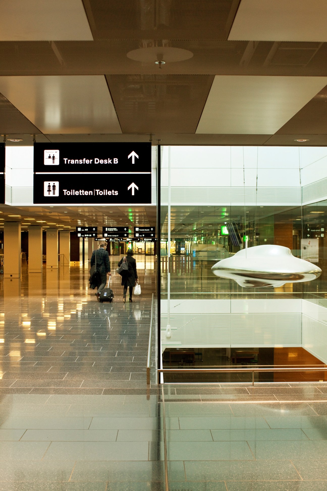 Modern airport terminal interior with directional signage and two travelers with luggage walking through polished corridors.