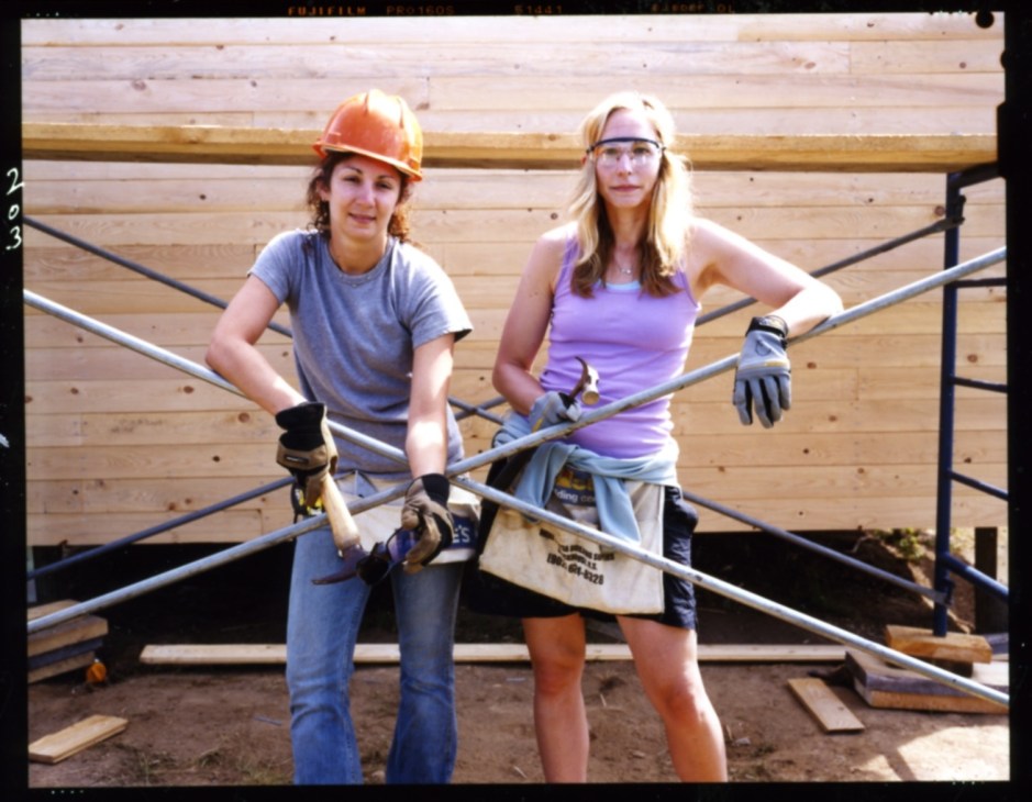 Two women wearing hard hats and work gloves stand on a wooden building frame under construction.