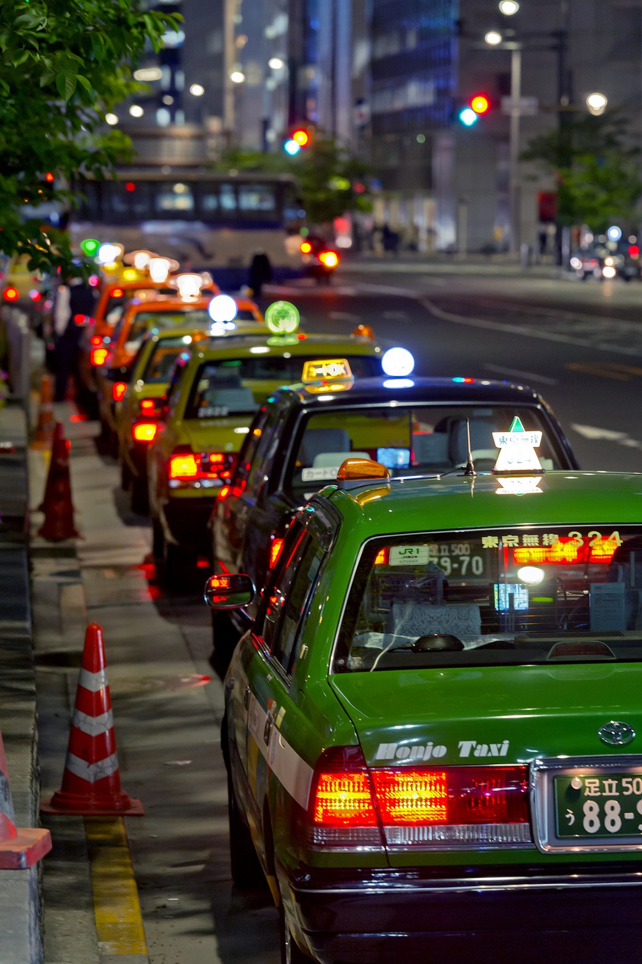 A queue of green and yellow Tokyo taxis lined up at night with illuminated taxi signs and traffic lights visible.