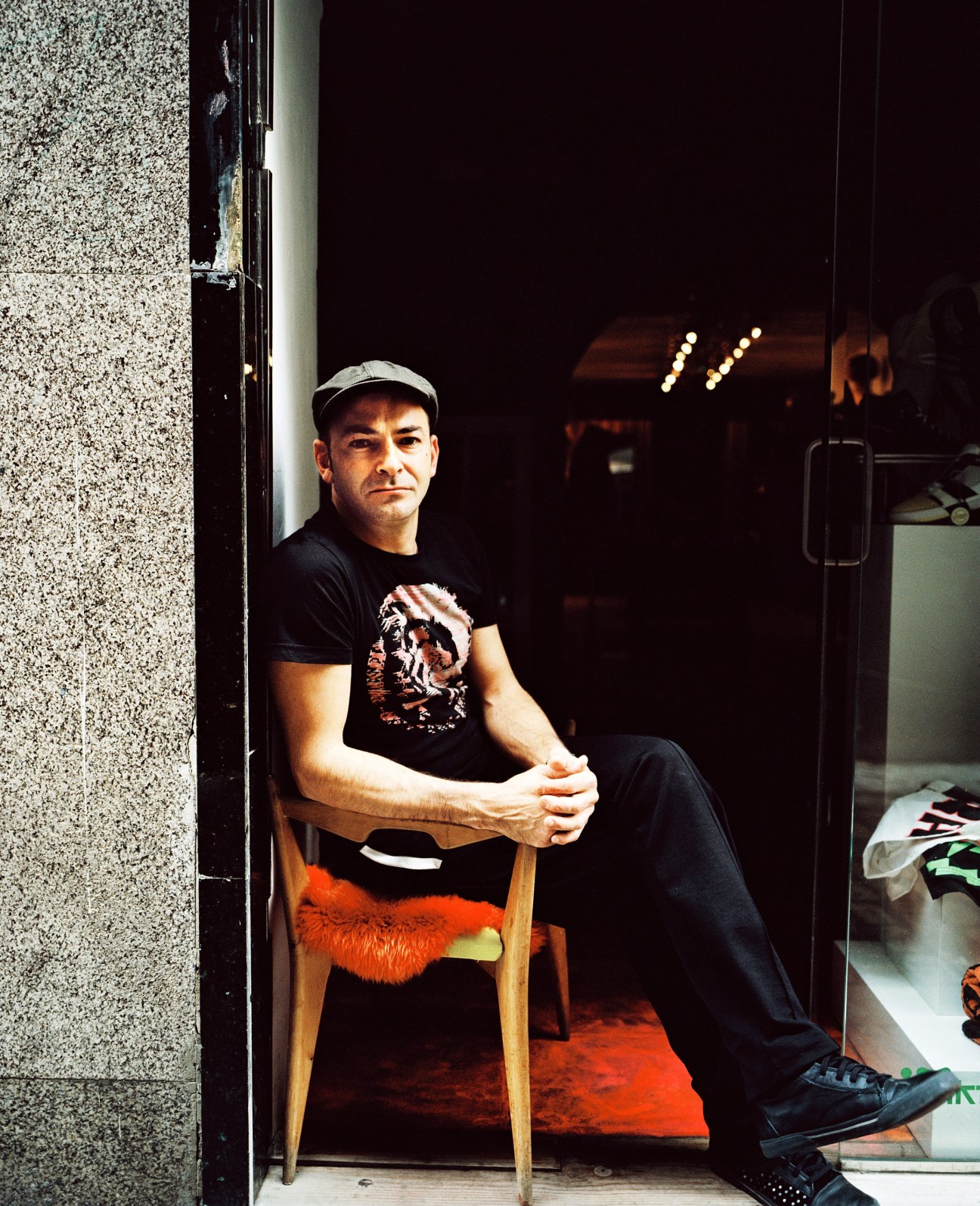 Arsenio Rodríguez Sánchez, owner of Arsenico boutique, sitting in a doorway wearing a black graphic t-shirt and cap.