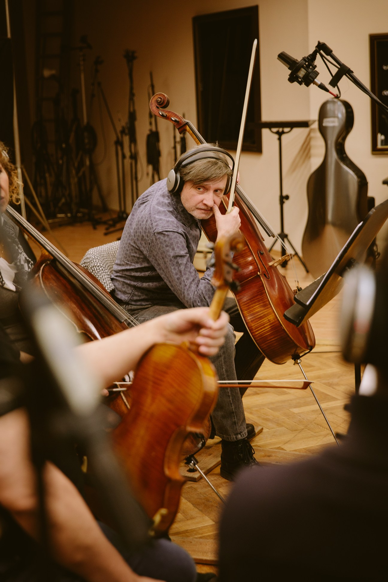Cellist Milos Jahoda wearing headphones plays in a recording studio surrounded by other musicians and microphone equipment.