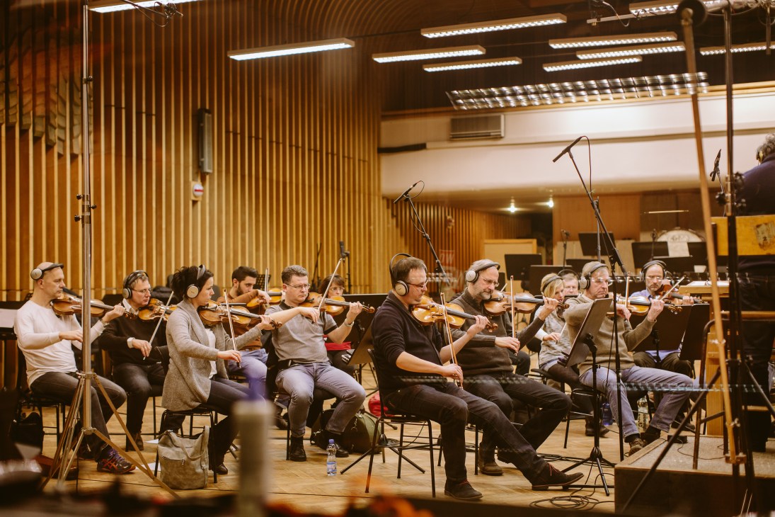 String musicians wearing headphones perform in a recording studio with wood paneling and overhead lighting.