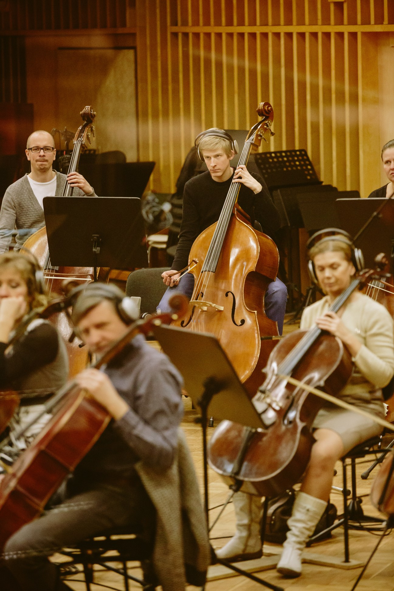 String musicians playing cellos in a recording studio with wood-paneled walls and music stands.