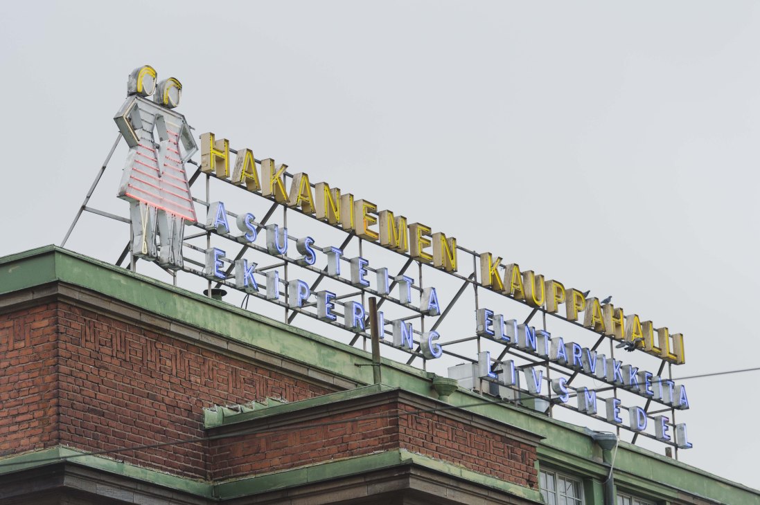 Neon sign for Hakaniemen Kauppahalli market hall in Helsinki with yellow and blue letters on a brick building.