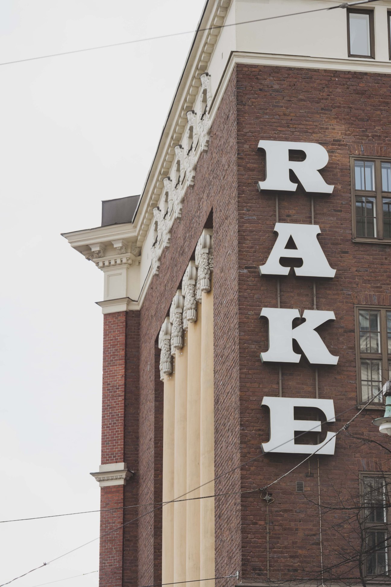White block letters spelling RAKE mounted vertically on a red brick building facade in Helsinki.