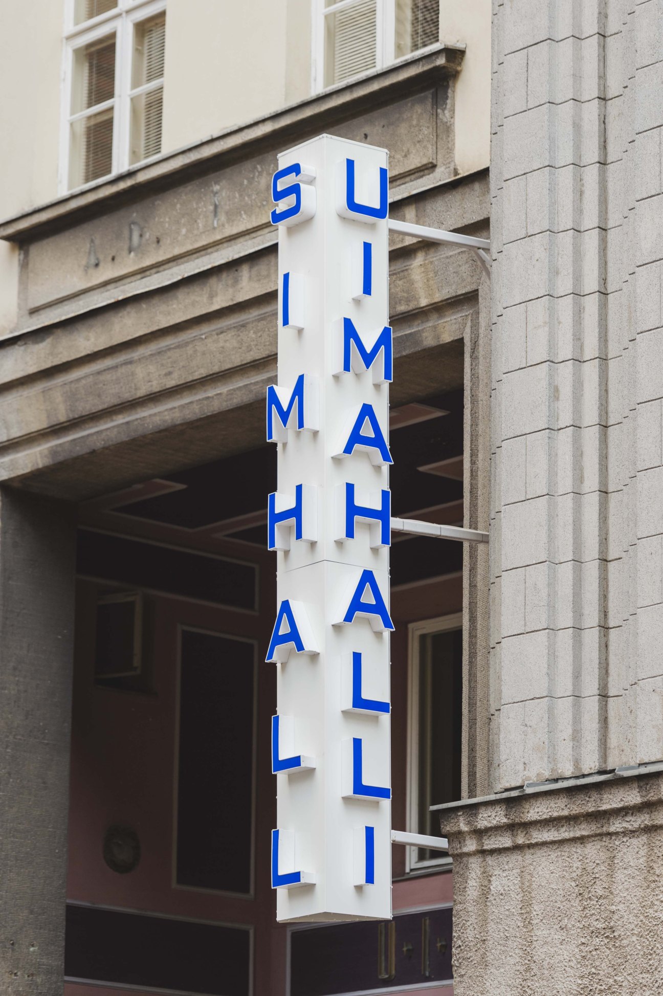 Vertical neon sign reading "SWIM HALL" with blue letters on a white background mounted on a building facade.