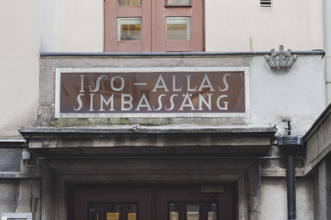 Brown storefront sign with white serif lettering reading 'ISSO-ALLAS SIMBASSANG' on a historic building facade.