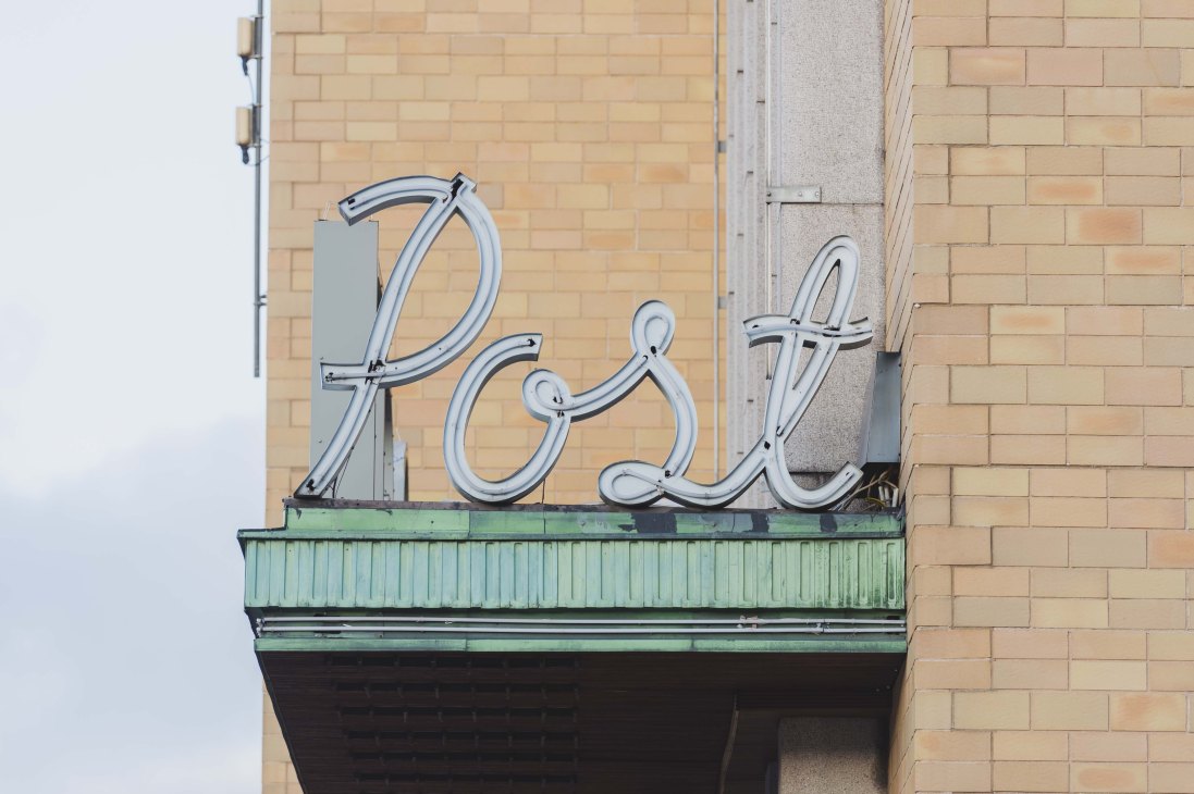 Vintage neon sign spelling 'Posti' in cursive script mounted on a brick building facade.