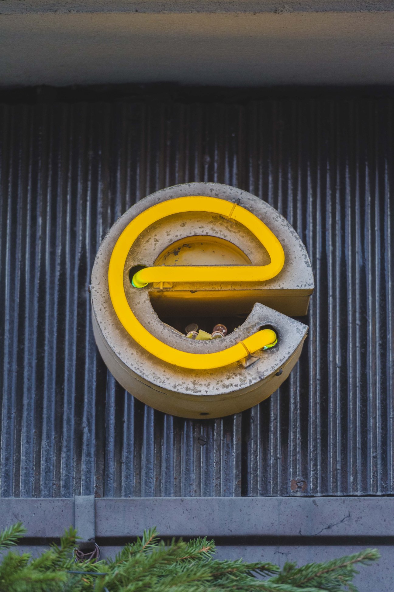 Yellow neon letter 'e' mounted on concrete backing against corrugated metal wall