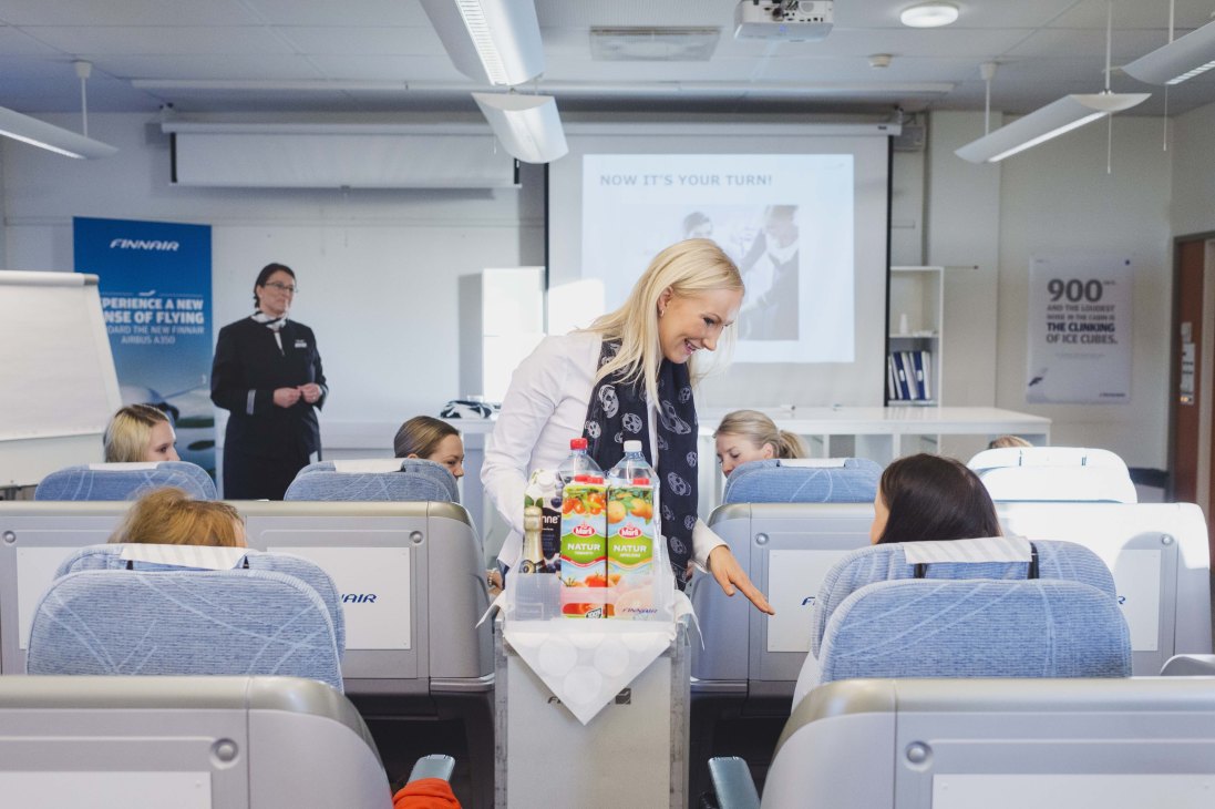 Finnair flight attendant serving beverages to students seated in Business Class replica seats during training.