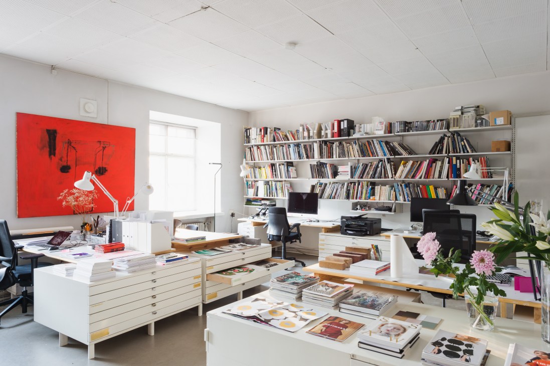 Creative studio workspace with white desk, red artwork, floor-to-ceiling bookshelves, and design materials.