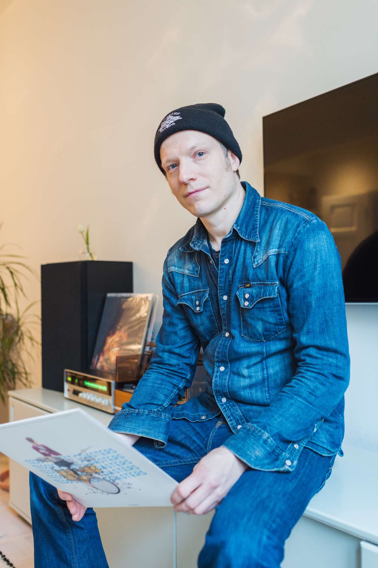 Musician Markus Nordenstreng in his home, wearing a denim shirt and black beanie, holding artwork.