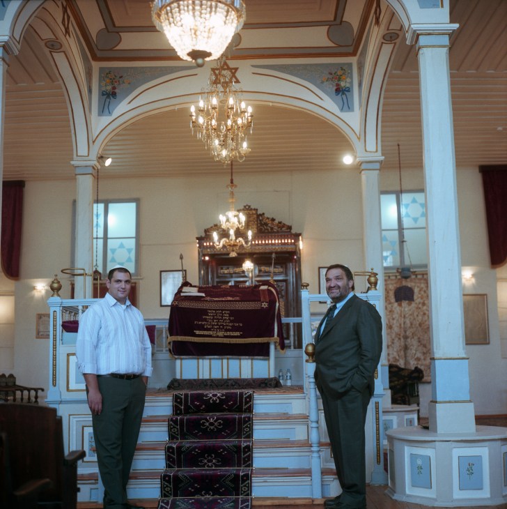 Two men stand in an ornate synagogue interior with chandeliers and blue columns.