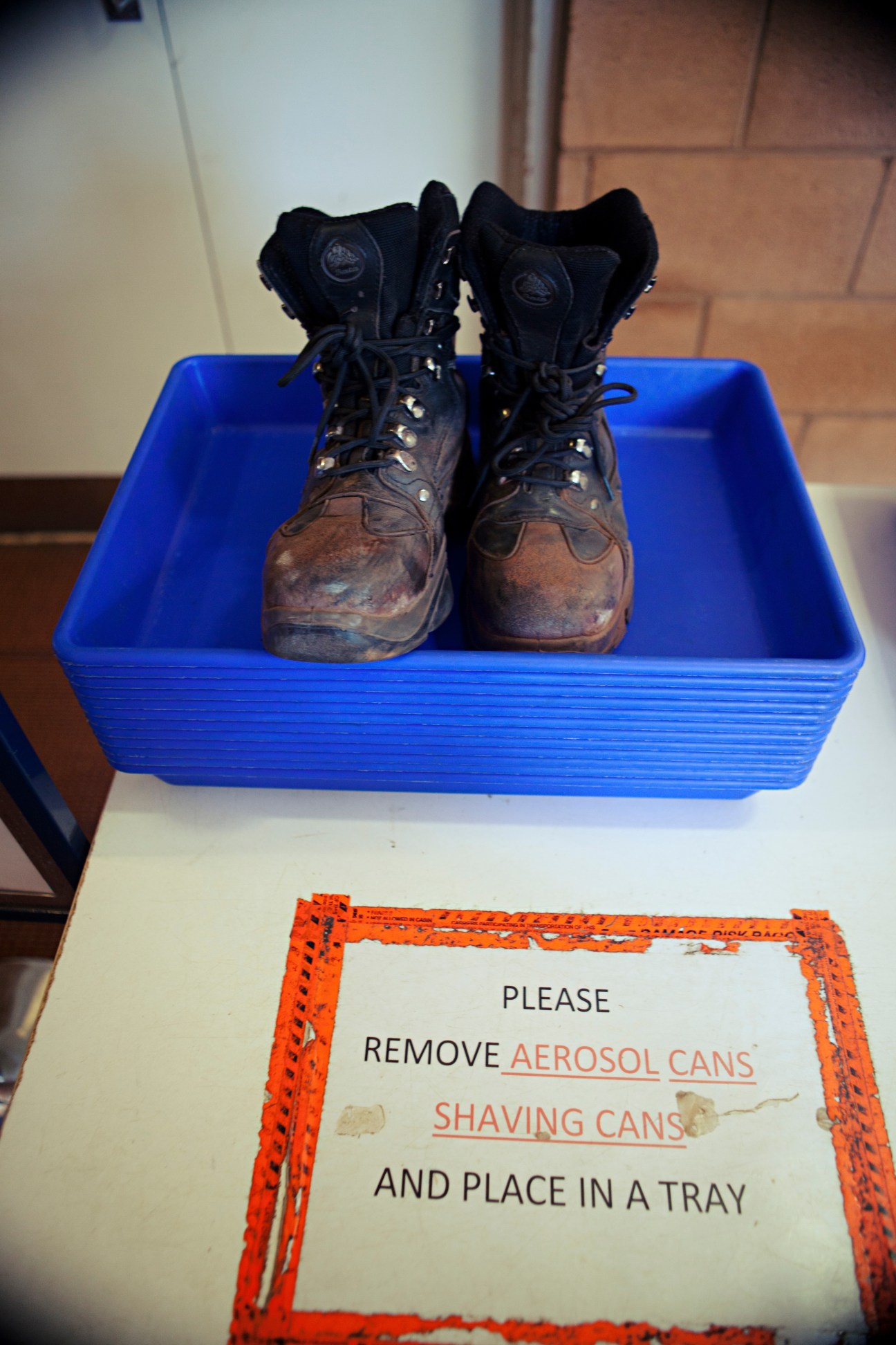 Dirty work boots in a blue tray at airport security with instructions for removing aerosol and shaving cans.