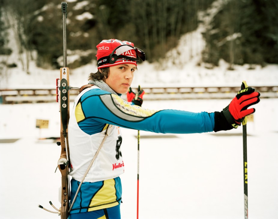 Military biathlon contestant in blue and yellow uniform holding rifle at snowy ski resort.
