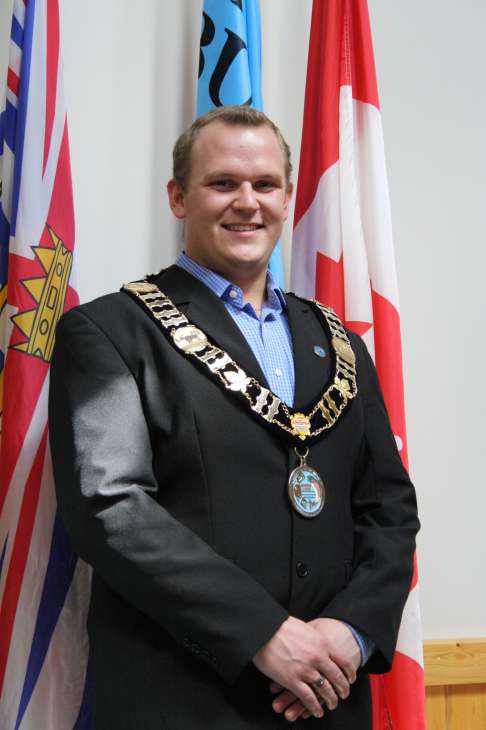 Luke Strimbold, Mayor of Burns Lake, wearing formal attire with an official chain of office and medals standing before Canadian flags.
