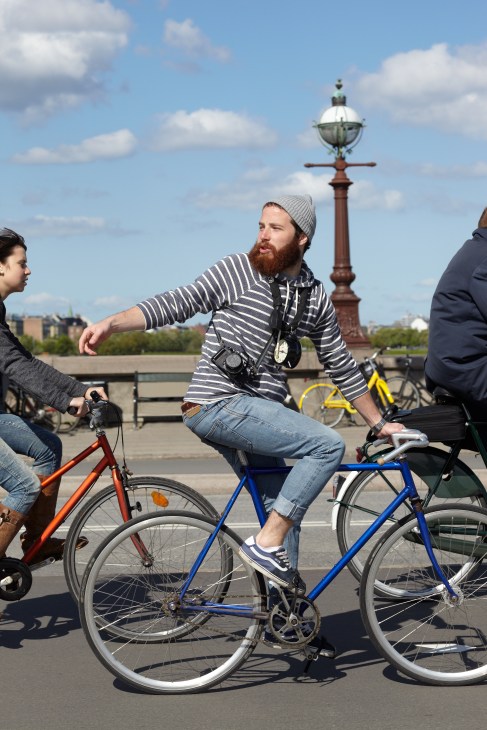 A man on a blue bicycle talks with two others on a bridge overlooking Copenhagen's waterfront and landmarks.