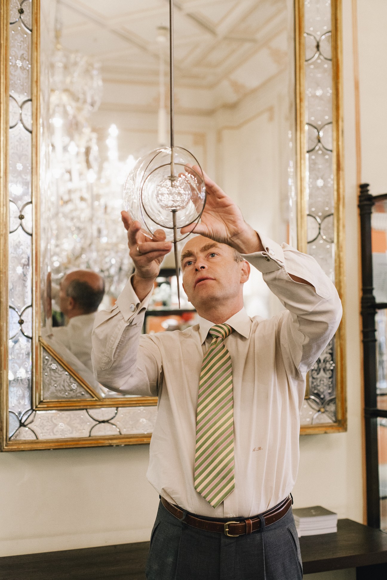 Man in white shirt and striped tie examines a crystal glass pendant in an ornate mirrored showroom.