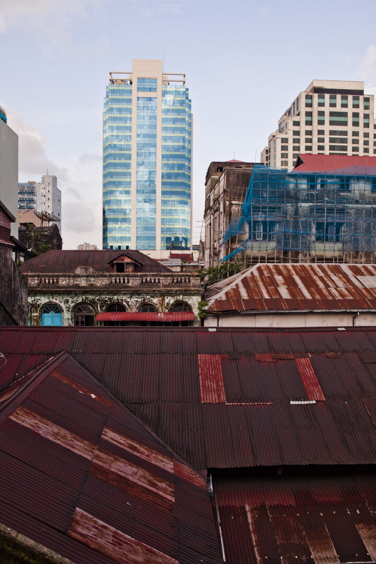 Decaying colonial building with blue-shuttered windows surrounded by modern high-rises and corrugated metal roofs in Rangoon.