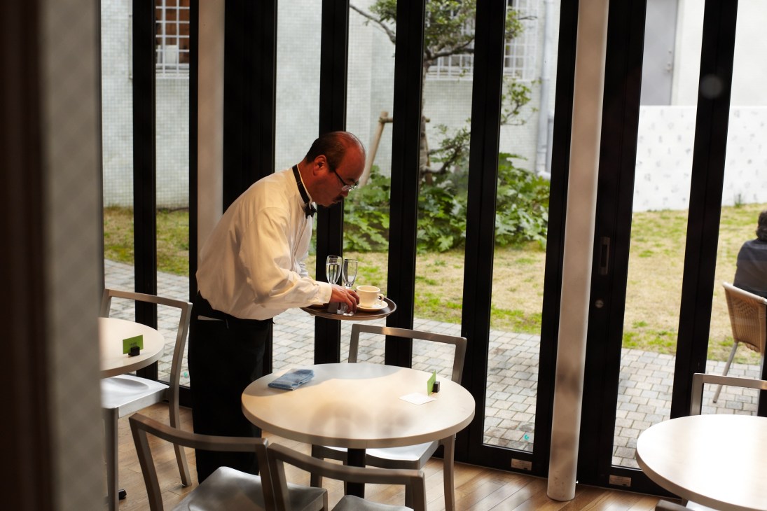 Server carrying drinks at a modern café with floor-to-ceiling black-framed windows overlooking a garden.