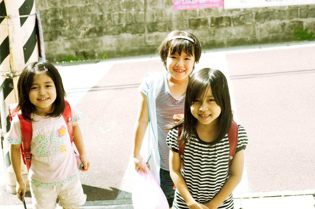 Three smiling primary school children standing together outdoors in Naha.