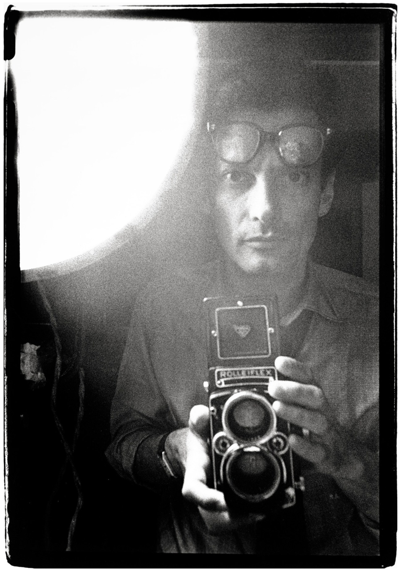 Black and white self-portrait of a man holding a medium format camera with glasses visible, taken in mirror reflection.