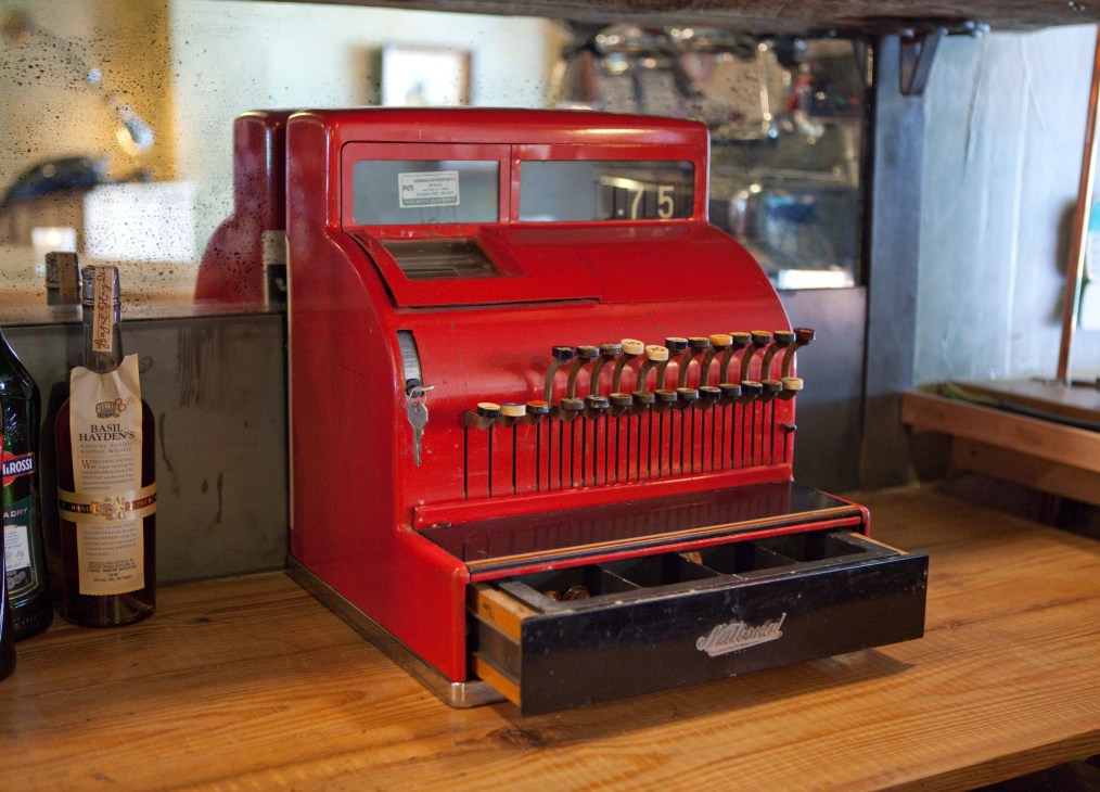 Red vintage cash register with numbered keys displayed on a wooden counter next to bottles.