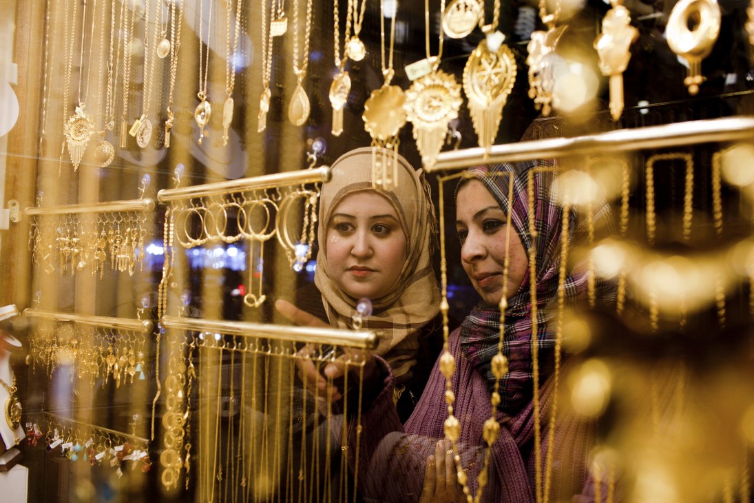 Two women wearing headscarves browse gold jewelry displayed in a shop window.