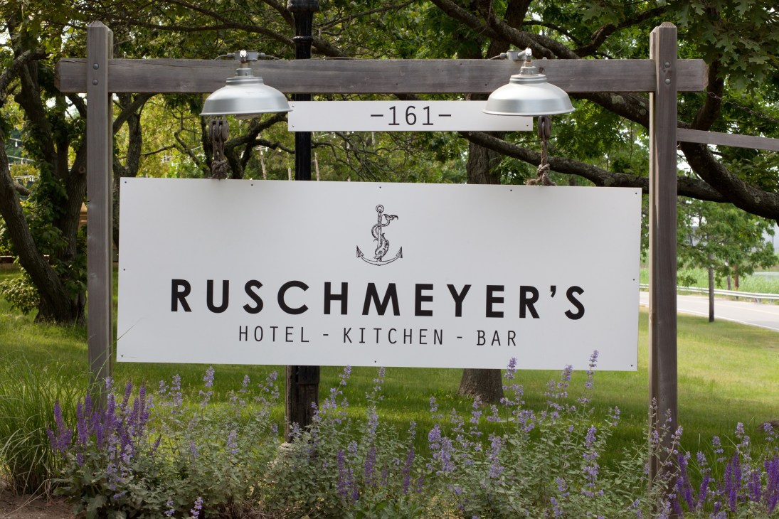 Ruschmeyer's hotel signage with anchor logo mounted on wooden frame surrounded by purple flowers.