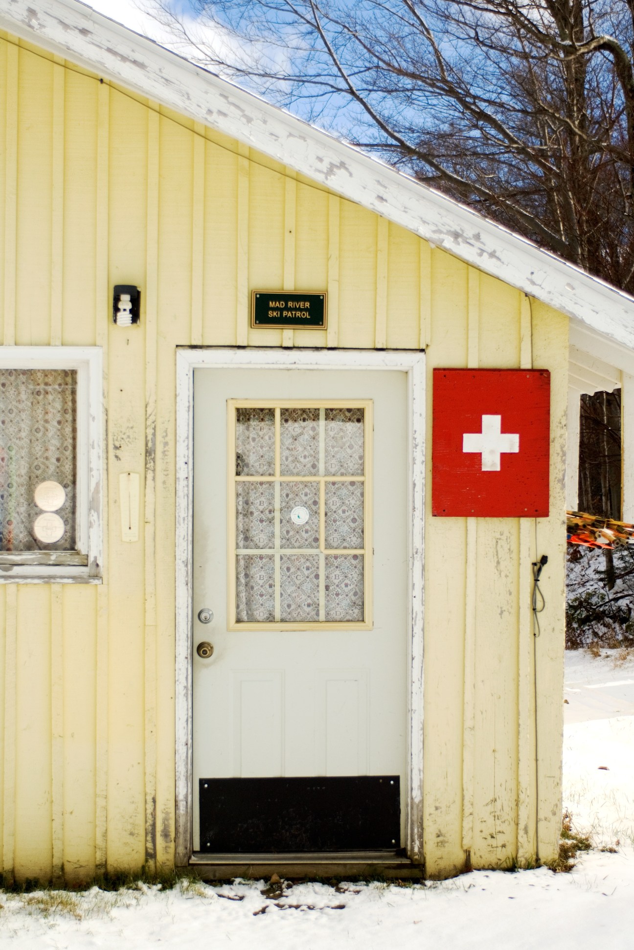 Yellow corrugated metal building with white door, red medical cross sign, and Mad River Ski Patrol plaque in snowy setting.