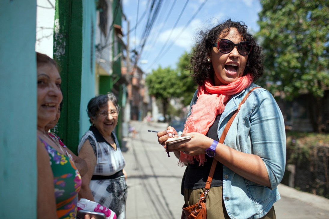Woman in denim shirt and red scarf gestures while speaking to residents on a colorful street lined with green buildings.