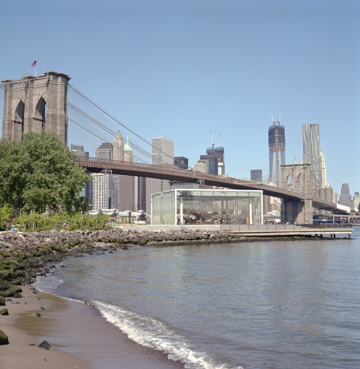 Jane's Carousel pavilion on Brooklyn waterfront with Brooklyn Bridge and Manhattan skyline beyond.