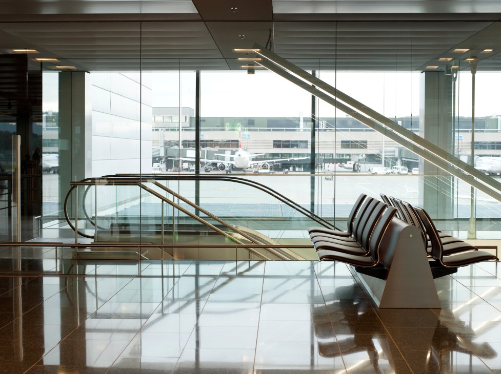 Modern airport terminal interior with curved staircase, floor-to-ceiling windows overlooking tarmac and aircraft.