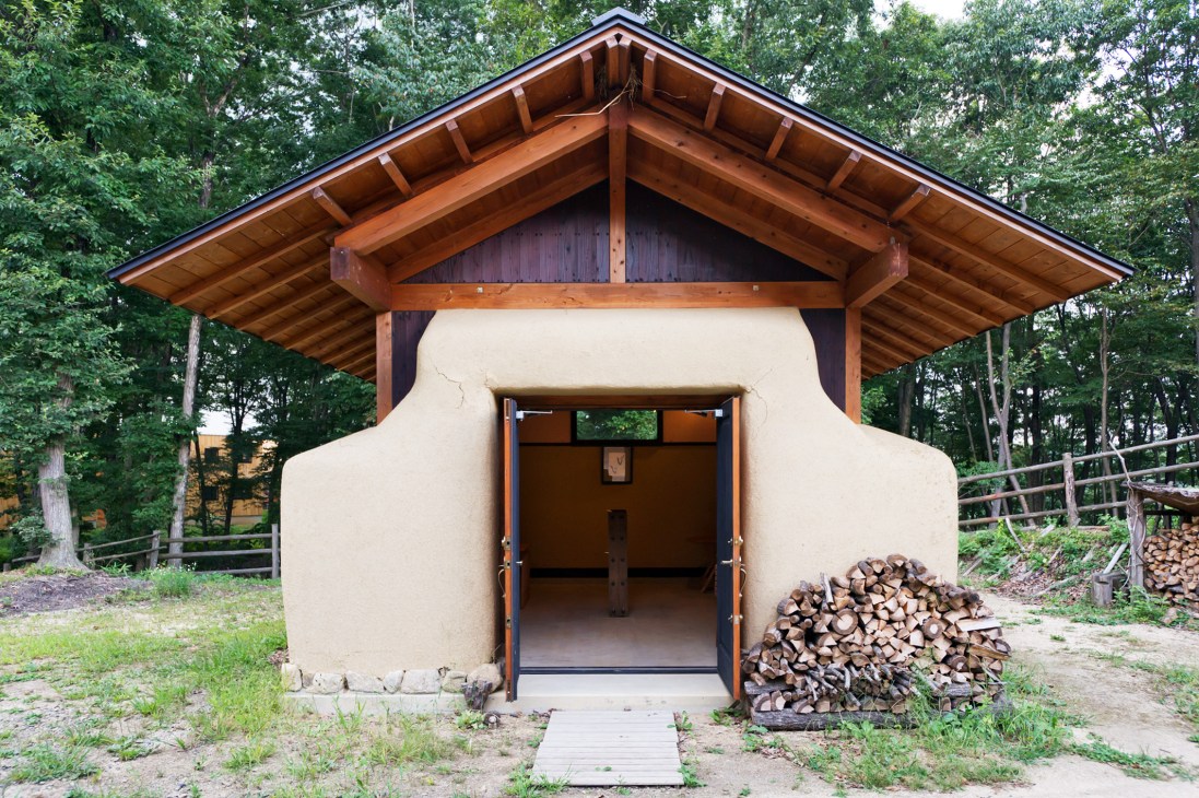 Modern building with curved cream walls, wooden roof frame, and open doorway surrounded by forest trees.