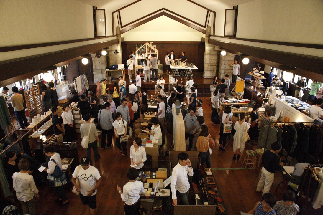 Overhead view of the For Stockists design and craft fair in Tokyo, showing numerous visitors browsing exhibitor stalls in a historic wooden interior space.