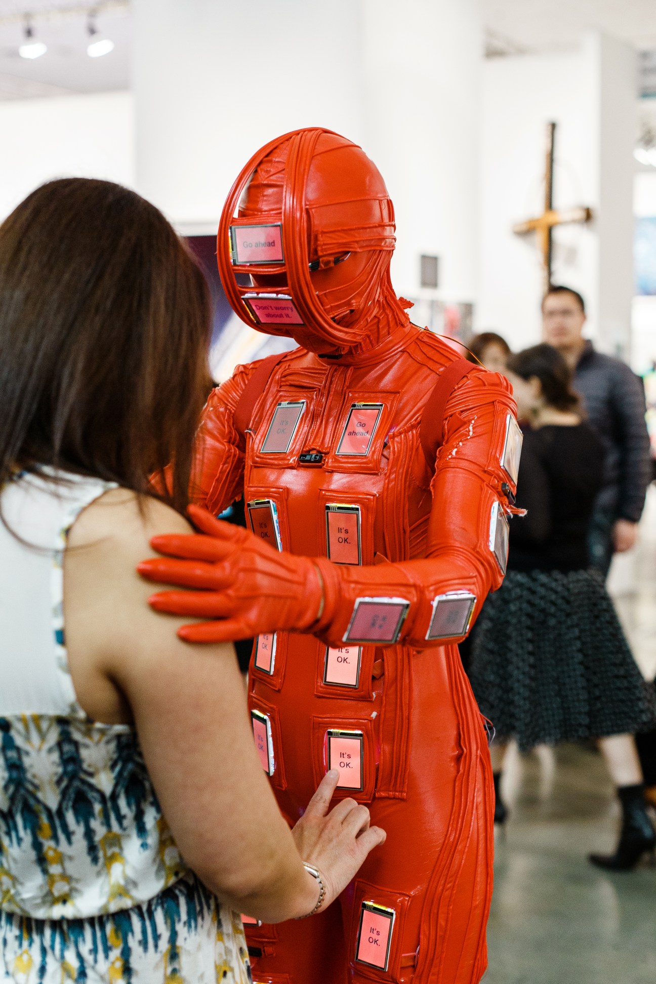 A person in a bright red interactive art suit covered with pink touchscreen buttons interacts with a gallery visitor.