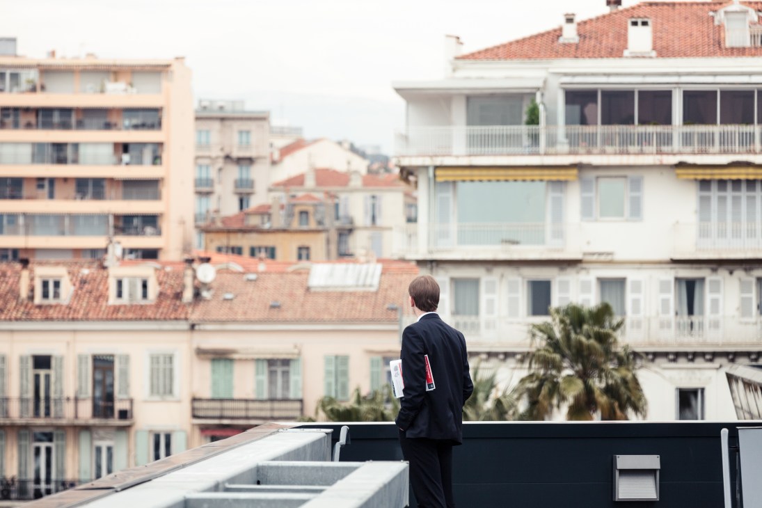 A man in a dark suit stands on a rooftop overlooking Cannes' Mediterranean architecture with terracotta roofs and palm trees.