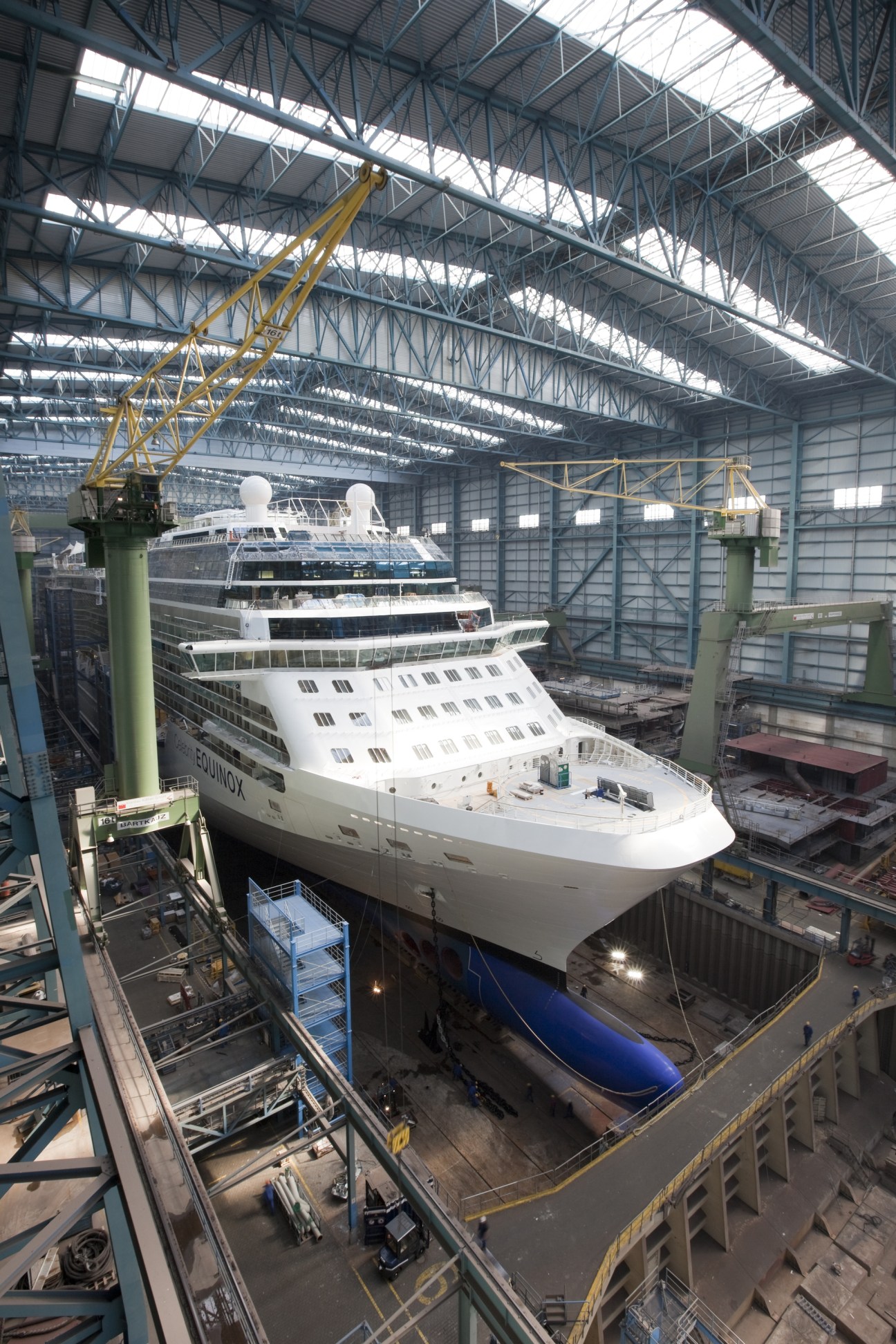 Large cruise ship under construction in a massive industrial shipyard with yellow cranes and steel framework overhead.