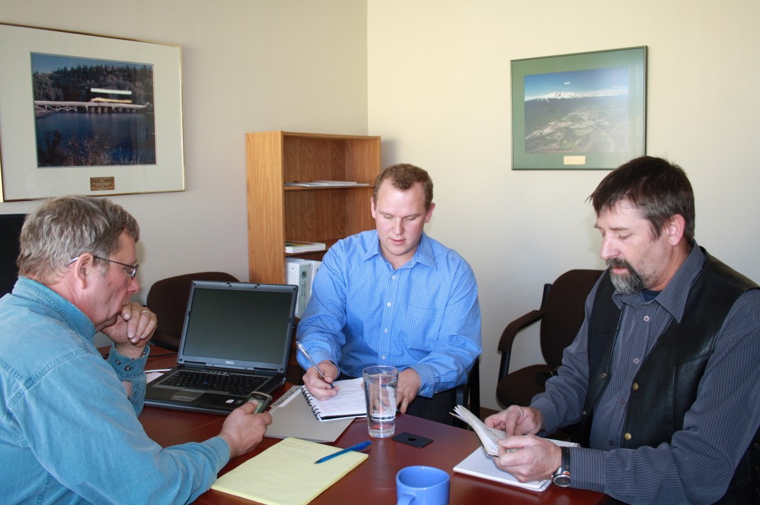 Three men in an office meeting with laptops and documents discussing local governance initiatives.