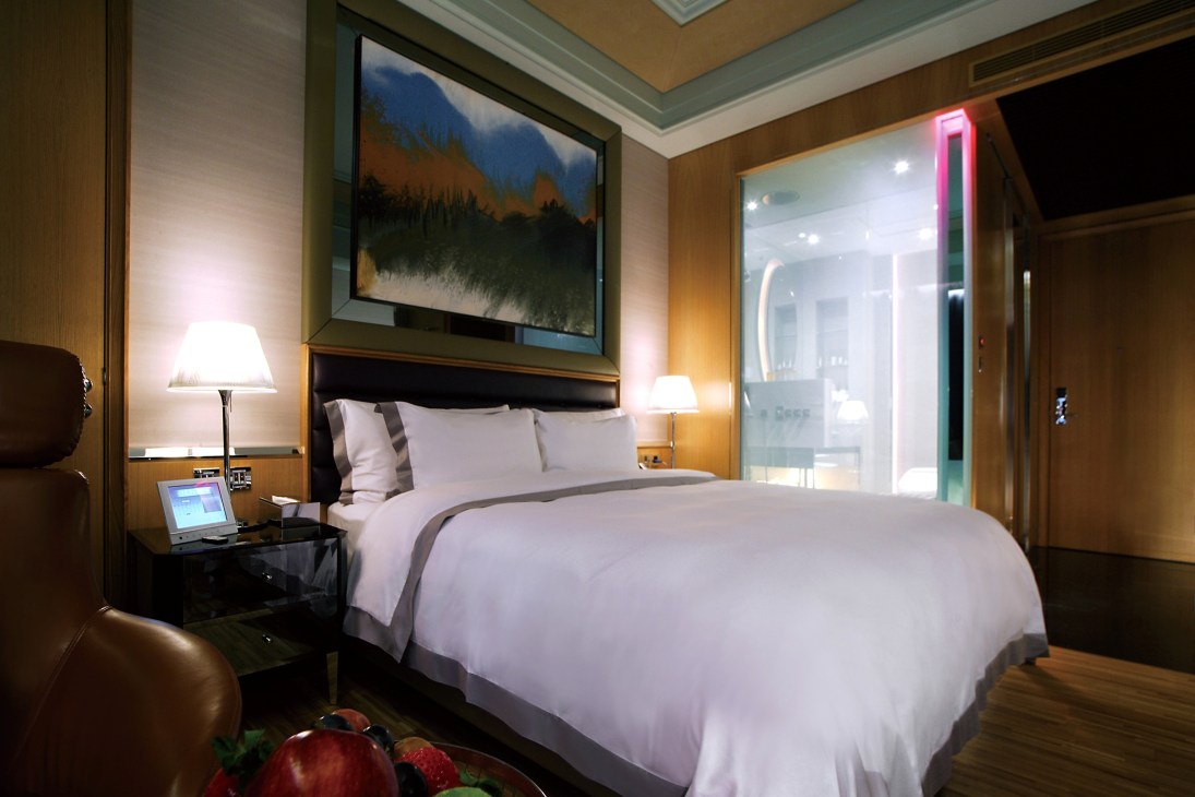 A modern hotel bedroom with warm wood paneling, a bed with white linens, bedside lamps, and a framed landscape artwork above the headboard.
