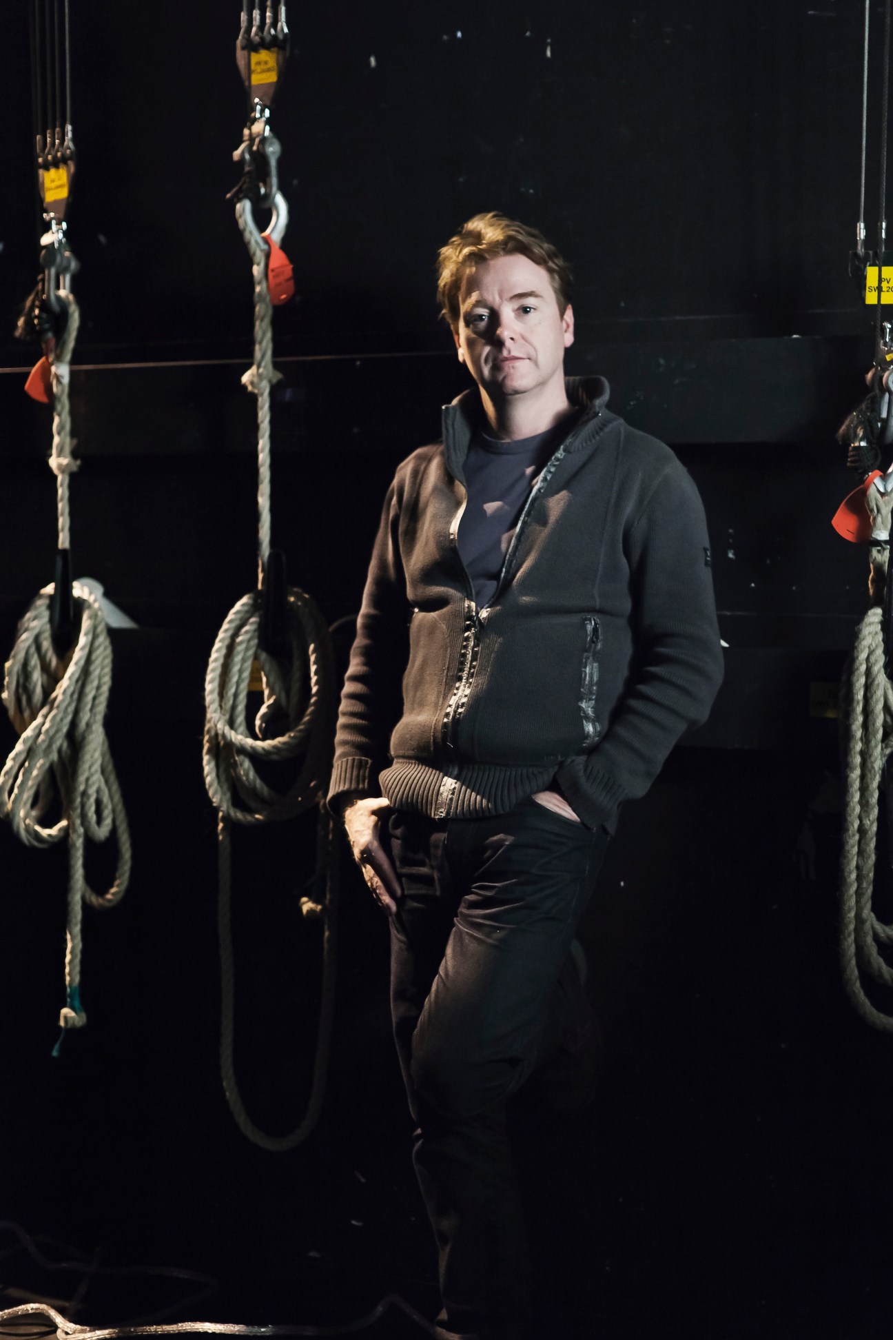 David McWilliams stands in front of maritime rigging and ropes against a dark background.