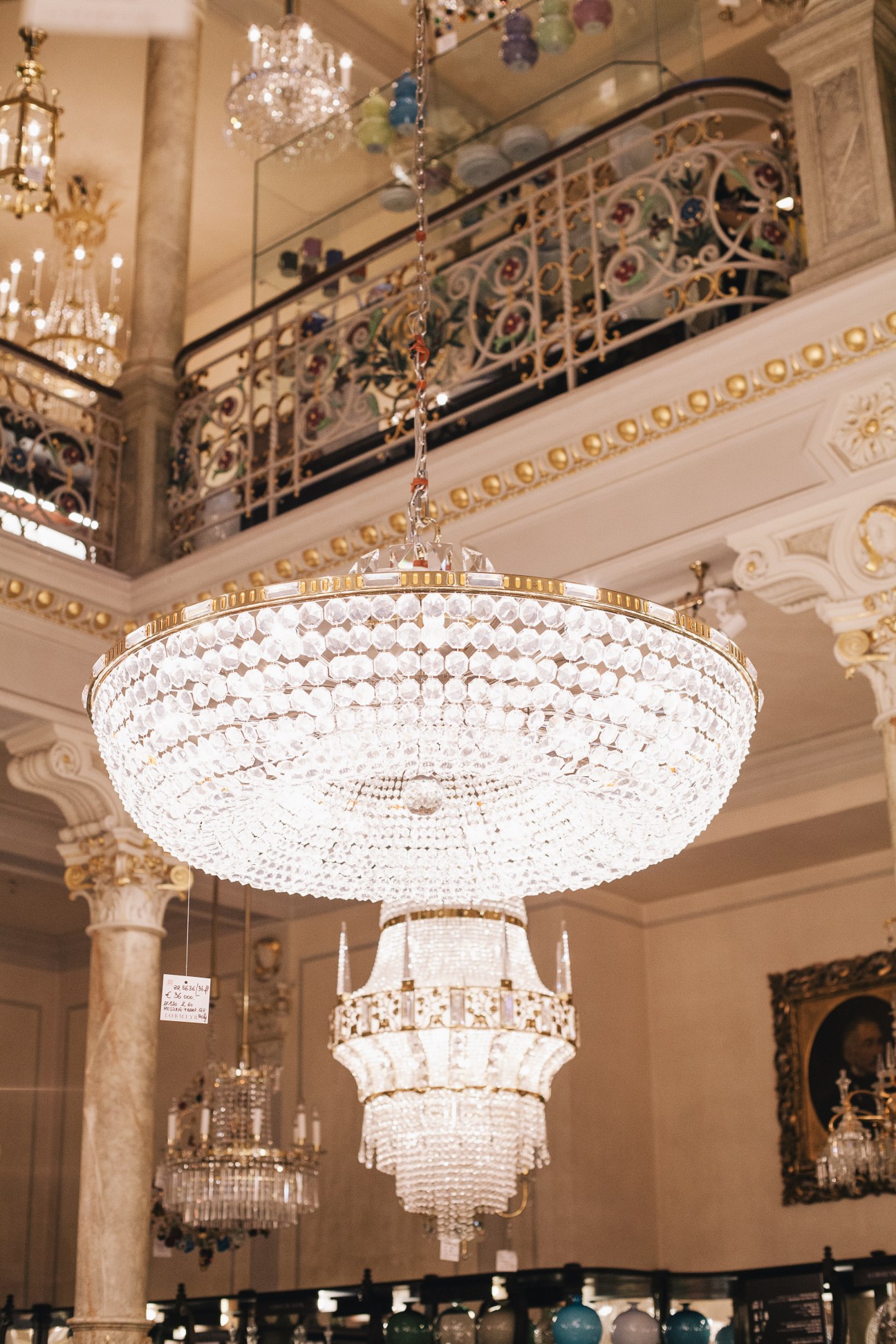 Crystal chandelier hanging in an ornate interior with decorative railings and architectural details.